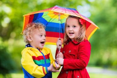 Little boy and girl play in rainy summer park. Children with colorful rainbow umbrella, waterproof jacket and coat playing in the rain. Kids walk in autumn shower. Outdoor fun by any weather.